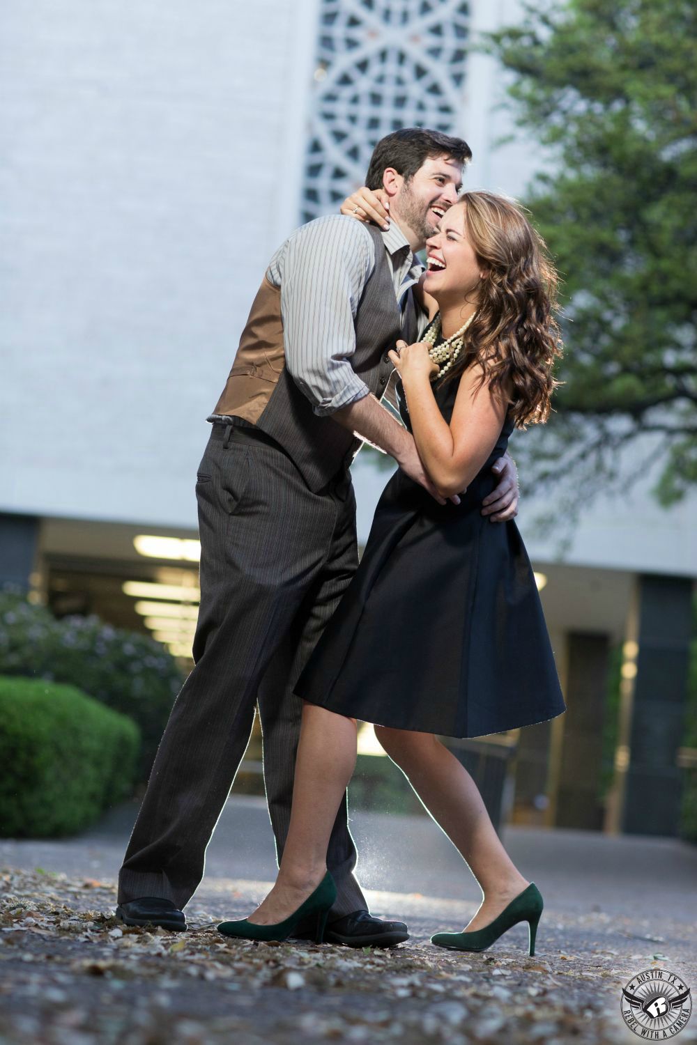 Brunette girl with an infectious laugh wearing black knee length dress, white pears and black heals holds a a guy with dark hair wearing a grey and black striped shirt, a tan and grey vest and grey striped pants in front of the library at UT in this sensational retro engagement image in Austin, Texas.